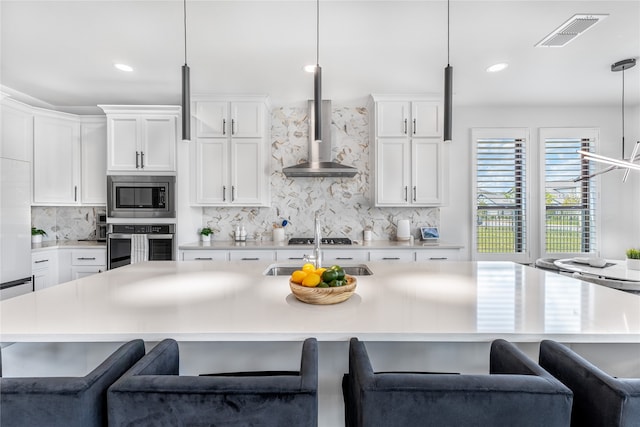 kitchen featuring hanging light fixtures, a kitchen island with sink, appliances with stainless steel finishes, white cabinets, and wall chimney exhaust hood