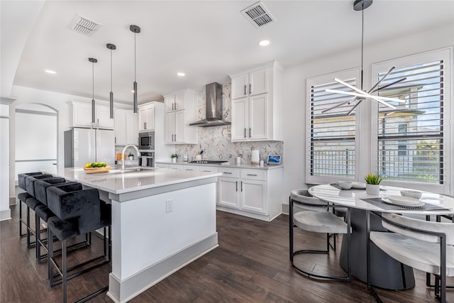 kitchen with white cabinetry, stainless steel appliances, a kitchen island with sink, hanging light fixtures, and wall chimney exhaust hood