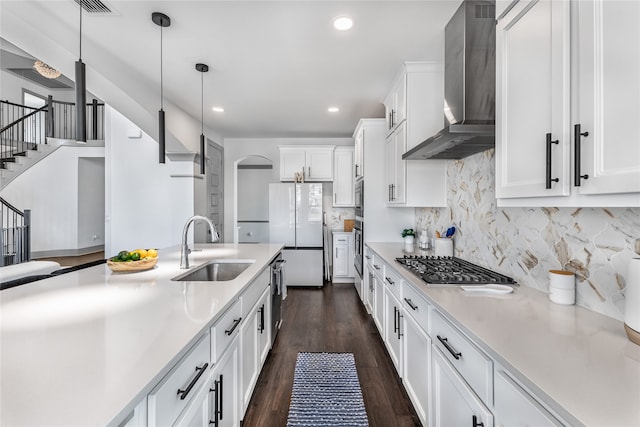 kitchen with white cabinets, wall chimney range hood, stainless steel appliances, sink, and hanging light fixtures