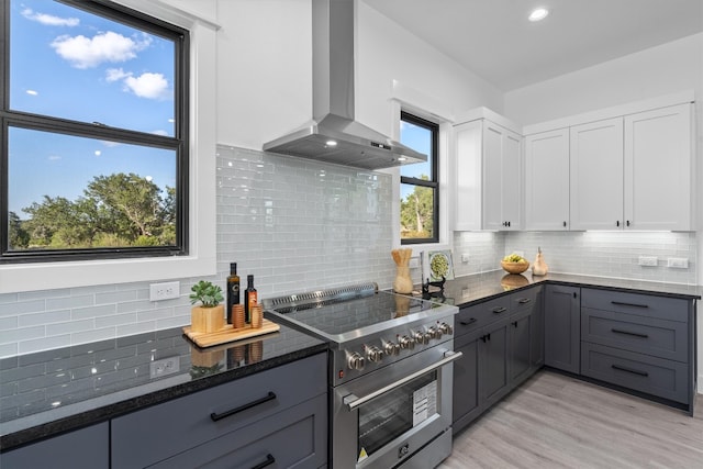 kitchen with double oven range, backsplash, dark stone counters, white cabinets, and wall chimney range hood