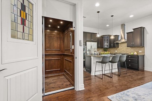 kitchen featuring dark wood-type flooring, an island with sink, wall chimney exhaust hood, and tasteful backsplash