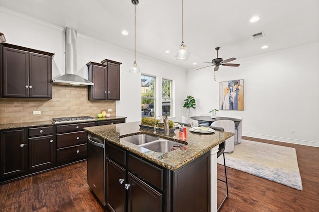 kitchen with dark wood-type flooring, decorative backsplash, an island with sink, ceiling fan, and wall chimney range hood