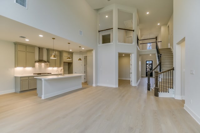 kitchen with a high ceiling, wall chimney exhaust hood, light hardwood / wood-style flooring, and a kitchen island with sink