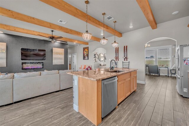 kitchen featuring beam ceiling, an island with sink, stainless steel appliances, sink, and ceiling fan