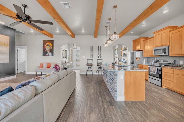 kitchen with beamed ceiling, dark hardwood / wood-style flooring, ceiling fan, appliances with stainless steel finishes, and a center island with sink