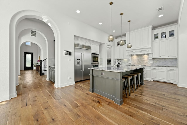 kitchen featuring light stone countertops, white cabinetry, built in appliances, tasteful backsplash, and an island with sink