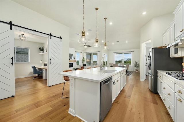 kitchen with appliances with stainless steel finishes, hanging light fixtures, an island with sink, white cabinets, and a barn door