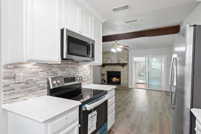 kitchen with a brick fireplace, appliances with stainless steel finishes, white cabinetry, beamed ceiling, and ceiling fan