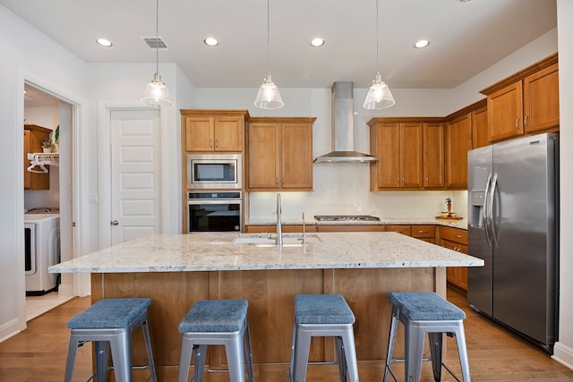 kitchen featuring stainless steel appliances, washer / dryer, a center island with sink, and wall chimney range hood