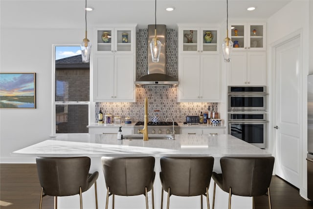 kitchen with pendant lighting, tasteful backsplash, stainless steel double oven, white cabinetry, and wall chimney range hood