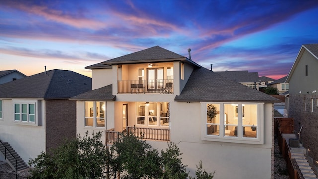 back of property at dusk featuring roof with shingles, stucco siding, a balcony, and a ceiling fan