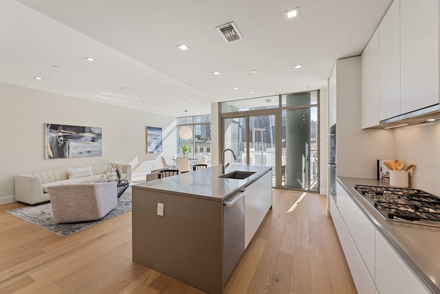 kitchen with floor to ceiling windows, white cabinets, sink, light hardwood / wood-style flooring, and an island with sink