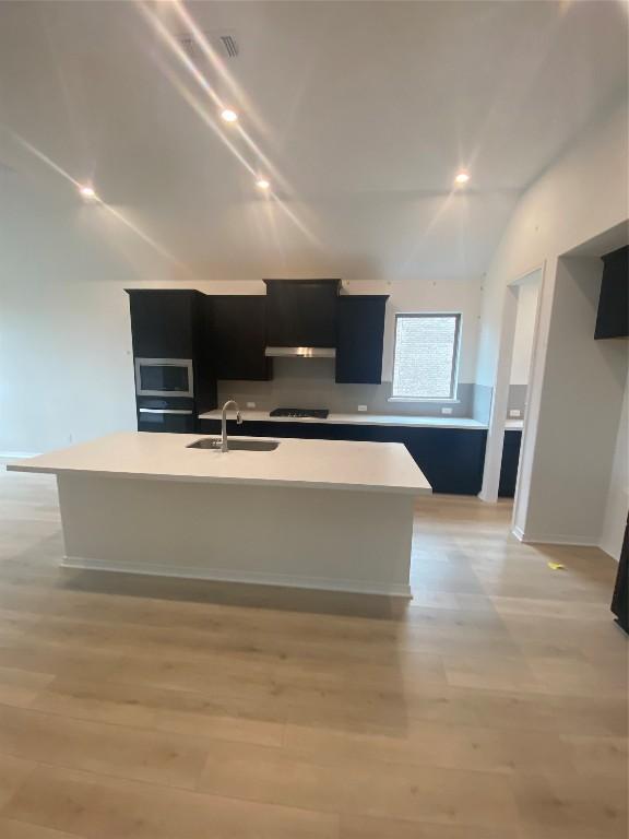 kitchen with vaulted ceiling, sink, black cooktop, a center island with sink, and light hardwood / wood-style flooring