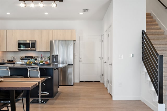 kitchen featuring light wood-type flooring, tasteful backsplash, light brown cabinetry, appliances with stainless steel finishes, and a breakfast bar area