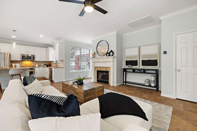 living room featuring ceiling fan, sink, crown molding, and a tiled fireplace