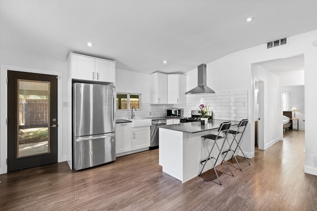 kitchen with stainless steel appliances, kitchen peninsula, dark hardwood / wood-style floors, white cabinets, and wall chimney range hood