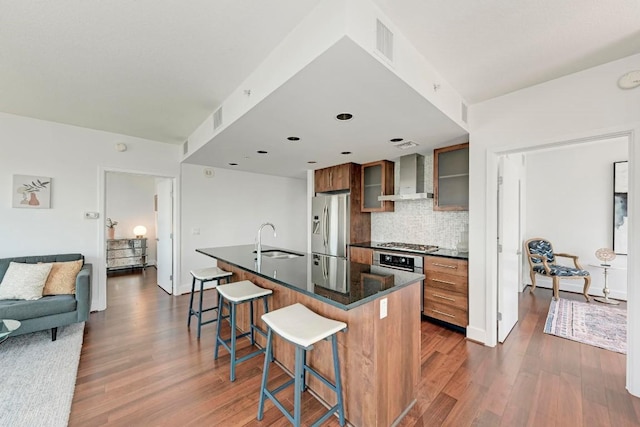 kitchen featuring wall chimney exhaust hood, dark countertops, appliances with stainless steel finishes, a sink, and a kitchen breakfast bar