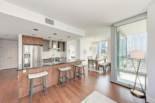 kitchen featuring stainless steel fridge with ice dispenser, wall chimney exhaust hood, tasteful backsplash, dark countertops, and a kitchen bar