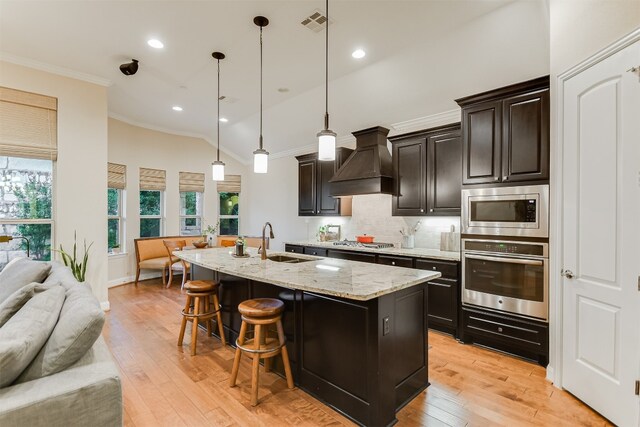 kitchen featuring custom exhaust hood, an island with sink, light hardwood / wood-style flooring, sink, and stainless steel appliances