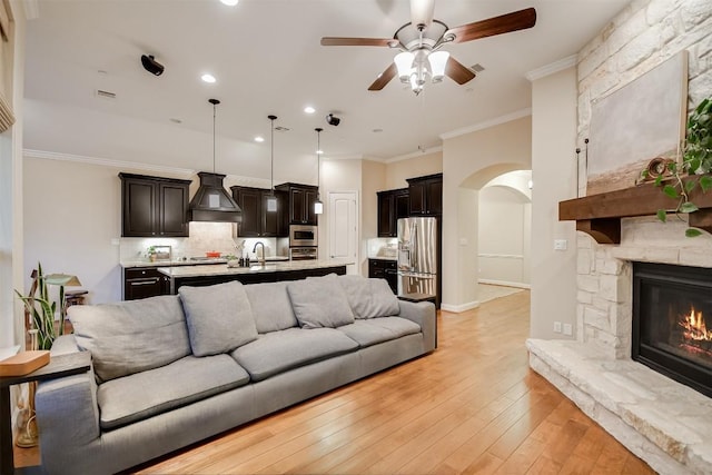 living area with arched walkways, a stone fireplace, light wood-style flooring, a ceiling fan, and crown molding