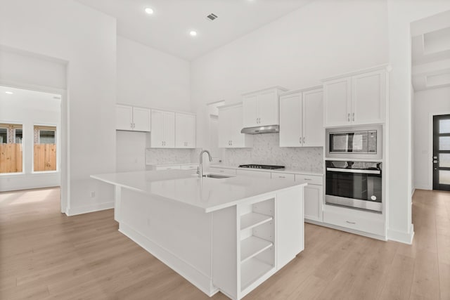 kitchen featuring a high ceiling, white cabinets, appliances with stainless steel finishes, light hardwood / wood-style floors, and a center island with sink