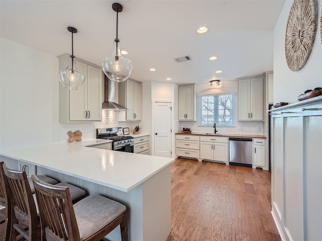 kitchen featuring hanging light fixtures, wall chimney range hood, kitchen peninsula, appliances with stainless steel finishes, and light wood-type flooring