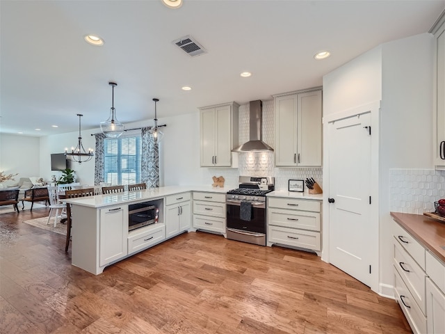 kitchen with kitchen peninsula, appliances with stainless steel finishes, wall chimney exhaust hood, light hardwood / wood-style floors, and hanging light fixtures