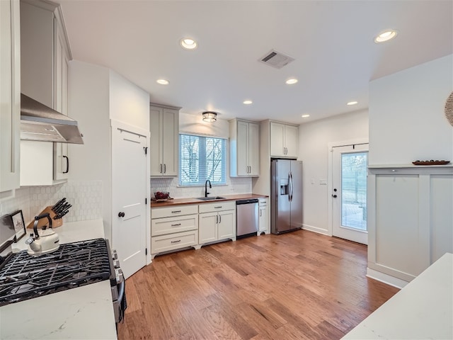 kitchen with appliances with stainless steel finishes, light wood-type flooring, backsplash, exhaust hood, and sink