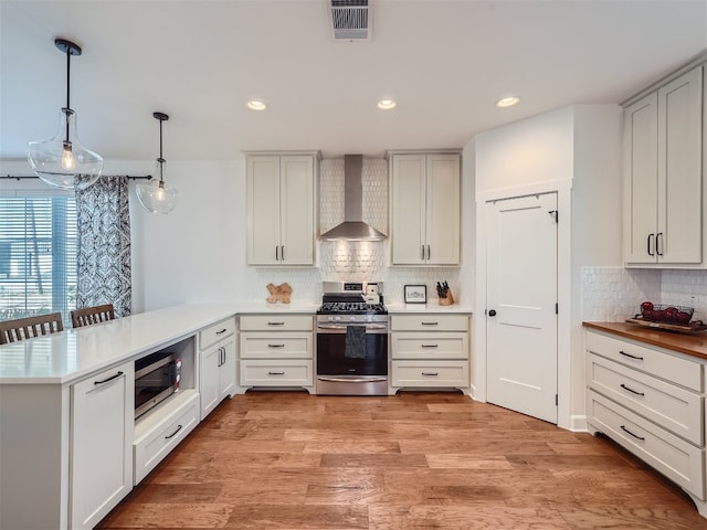 kitchen with stainless steel appliances, wall chimney range hood, kitchen peninsula, decorative light fixtures, and light wood-type flooring