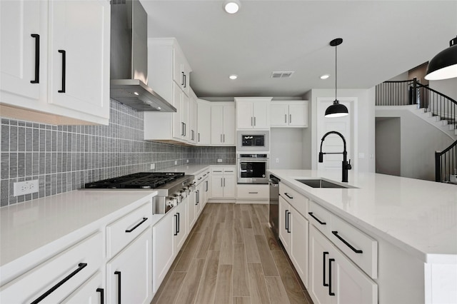 kitchen featuring sink, wall chimney exhaust hood, appliances with stainless steel finishes, decorative light fixtures, and white cabinetry