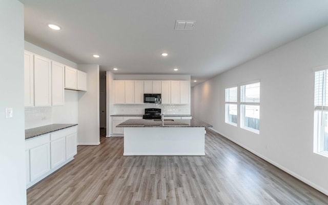 kitchen with light hardwood / wood-style flooring, dark stone countertops, a center island with sink, white cabinets, and black appliances