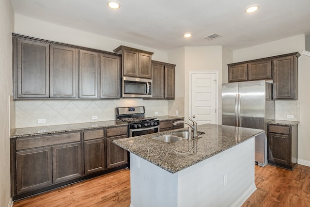 kitchen featuring appliances with stainless steel finishes, light hardwood / wood-style floors, sink, an island with sink, and dark stone counters