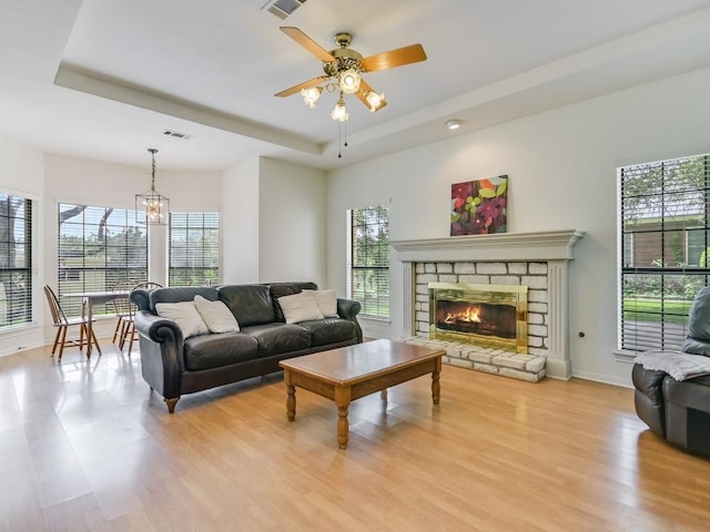 living area featuring visible vents, light wood-style flooring, a stone fireplace, a raised ceiling, and ceiling fan with notable chandelier