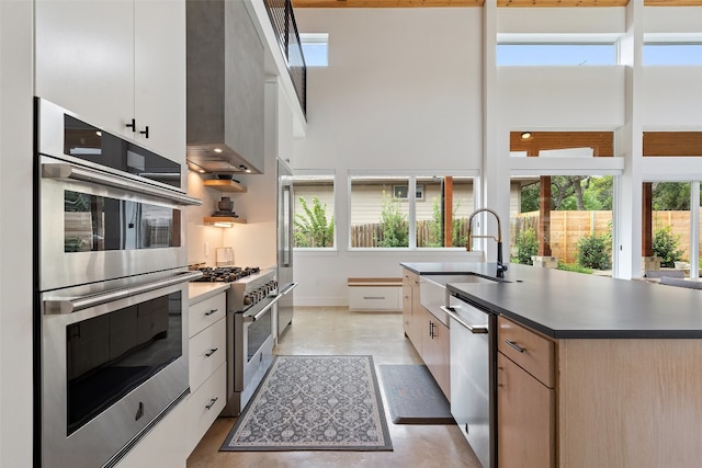 kitchen with high end appliances, an island with sink, sink, wall chimney range hood, and white cabinets