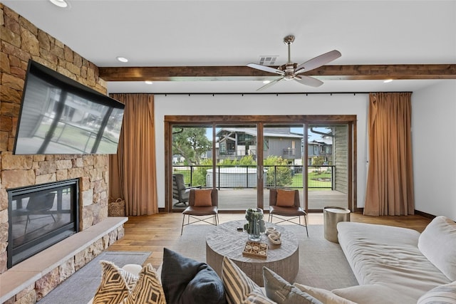 living room featuring light wood-type flooring, beamed ceiling, ceiling fan, and a stone fireplace