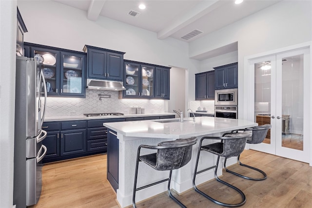 kitchen with light wood-type flooring, stainless steel appliances, a center island with sink, sink, and beam ceiling