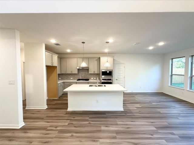 kitchen featuring appliances with stainless steel finishes, backsplash, a kitchen island with sink, wood-type flooring, and hanging light fixtures