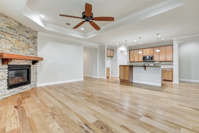 unfurnished living room featuring light hardwood / wood-style floors, a raised ceiling, a stone fireplace, crown molding, and ceiling fan