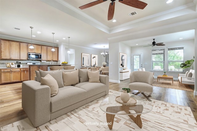 living room with light wood-type flooring, ceiling fan with notable chandelier, crown molding, and a tray ceiling