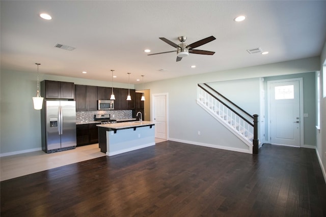 kitchen with dark brown cabinets, wood-type flooring, an island with sink, ceiling fan, and appliances with stainless steel finishes