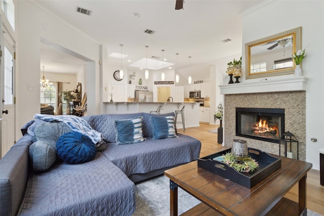 living room featuring crown molding, a tiled fireplace, ceiling fan with notable chandelier, and light wood-type flooring