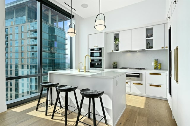 kitchen with stainless steel double oven, a kitchen island with sink, light hardwood / wood-style flooring, white cabinetry, and hanging light fixtures