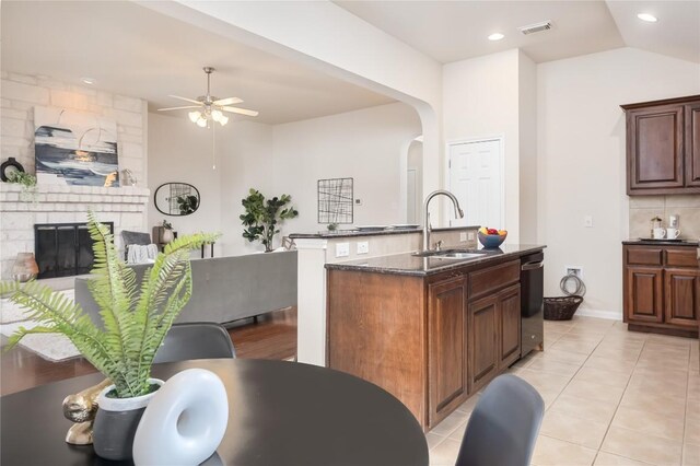 kitchen with sink, stainless steel dishwasher, dark stone counters, a fireplace, and a center island with sink