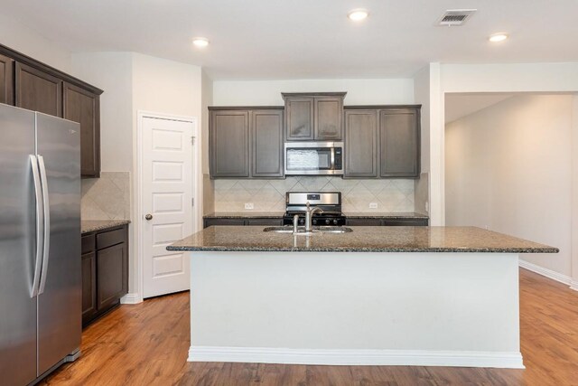 kitchen featuring an island with sink, stainless steel appliances, dark stone countertops, and light wood-type flooring