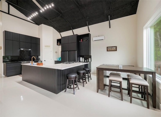 kitchen with a kitchen bar, a wall mounted AC, an island with sink, stainless steel gas stovetop, and backsplash