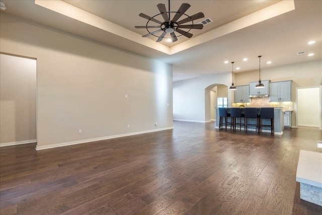 unfurnished living room with arched walkways, dark wood-type flooring, visible vents, a ceiling fan, and a raised ceiling