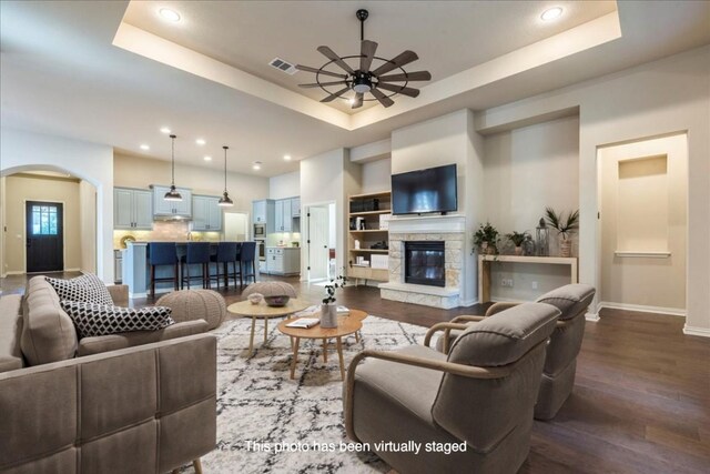 living room with ceiling fan, a stone fireplace, a raised ceiling, and wood-type flooring