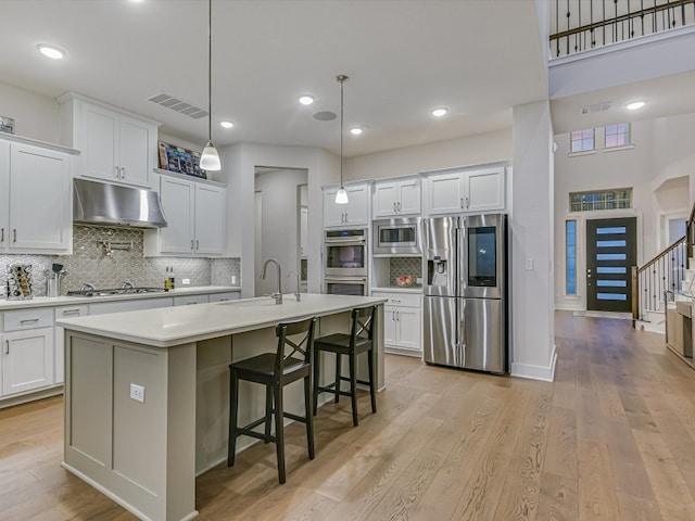 kitchen with hanging light fixtures, light wood-type flooring, stainless steel appliances, white cabinetry, and a center island with sink