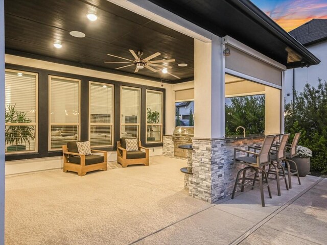 patio terrace at dusk with area for grilling, ceiling fan, a grill, and exterior bar