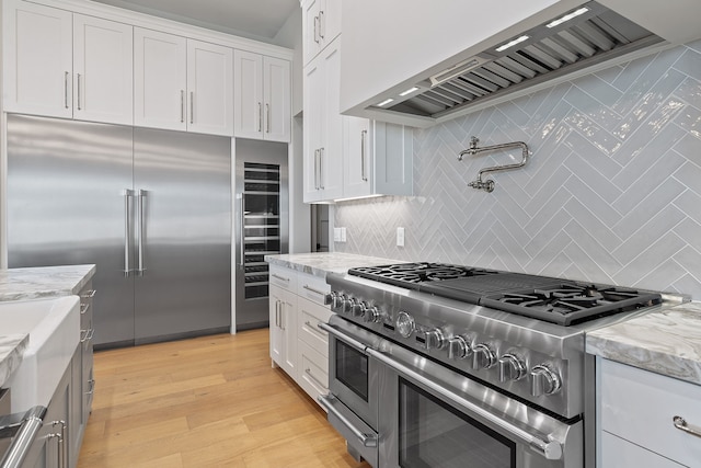 kitchen with custom exhaust hood, light stone countertops, high end appliances, white cabinets, and light wood-type flooring